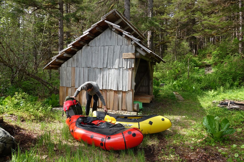 A man pulls up packrafts by a wooden three-sided shelter surrounded by trees on the Cross-Admiralty Island Canoe Route