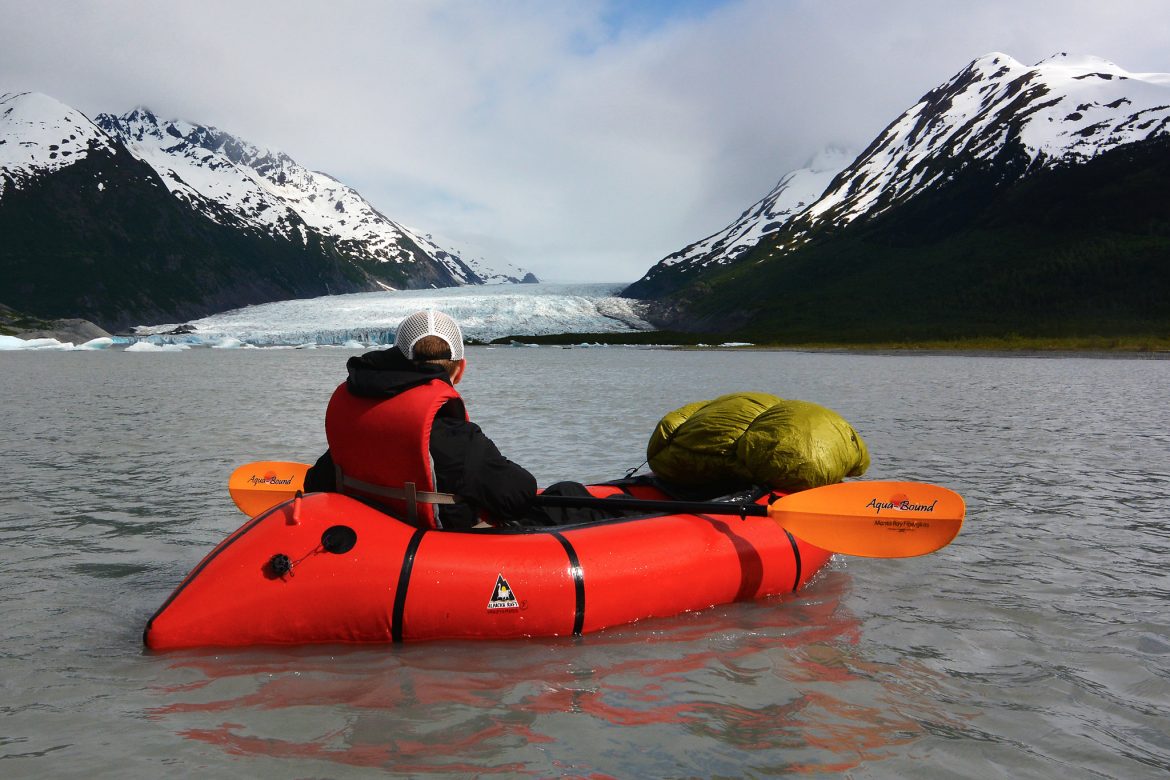 Paddler sits on lake in red packraft and looks at mountains and glacier