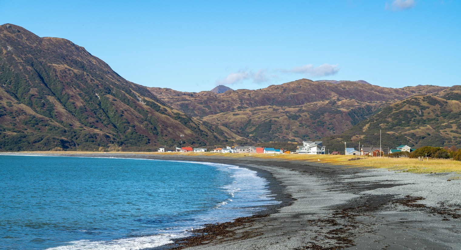 Blue ocean bay with small waves lapping at gravel beach and a small community nestled beneath mountains
