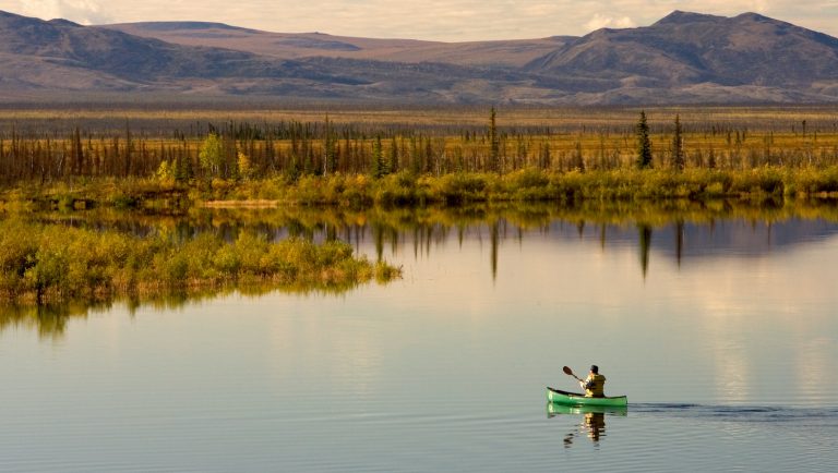 Man in green canoe paddles across calm lake with fall foliage and mountains ahead