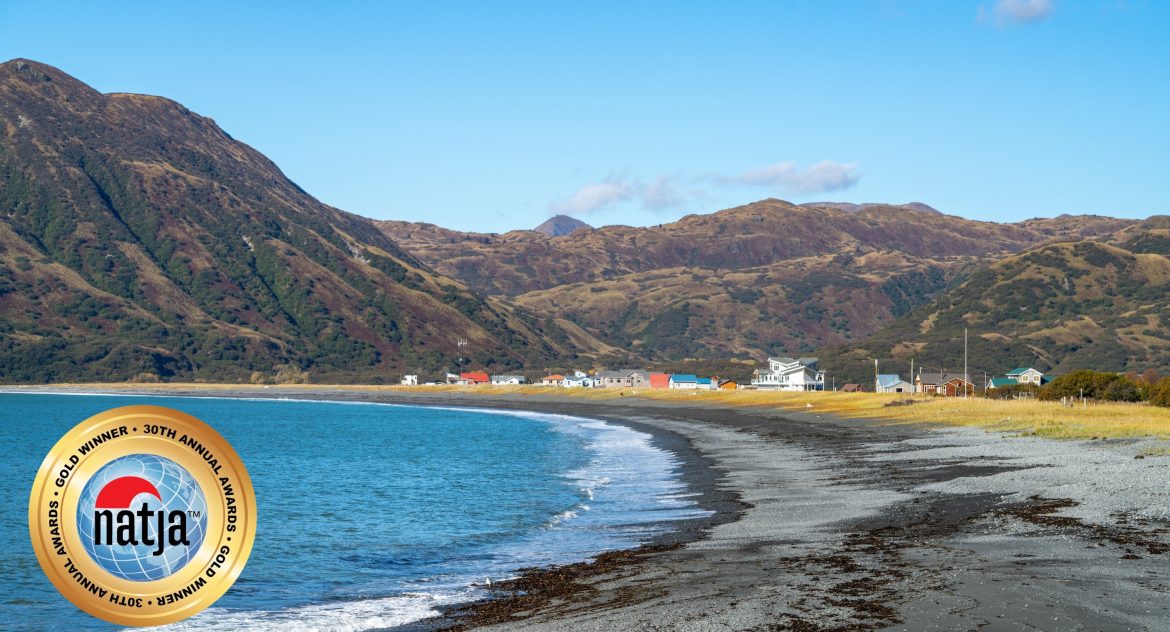 Blue ocean bay with small waves lapping at gravel beach and a small community nestled beneath mountains