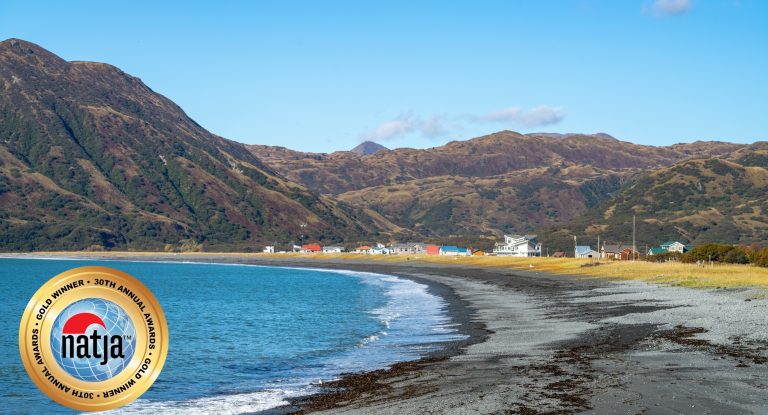 Blue ocean bay with small waves lapping at gravel beach and a small community nestled beneath mountains