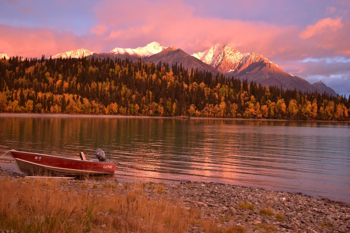 Skiff rests on shore of a calm lake, mountains, and pink clouds in backgrounds