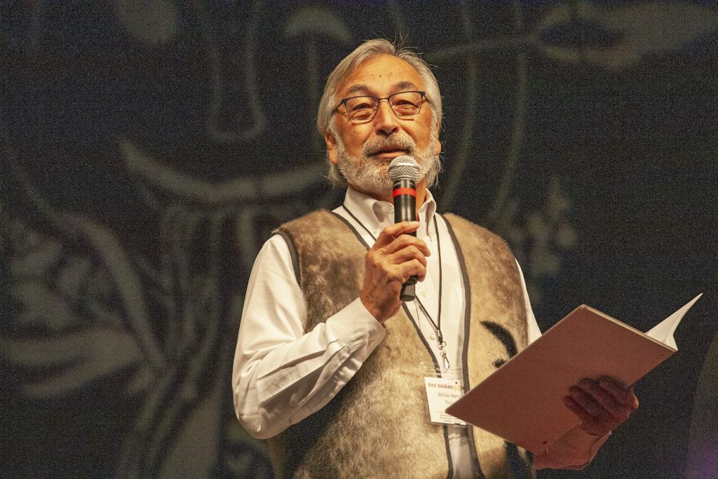 Gray-haired Alaska Native man with beard wearing a vest speaking on microphone