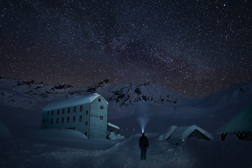 Man stands silhouetted against snow covered landscape, old Independence Mine Building behind him, and a sky full of stars. Man's headlamp lights a cone of the air above him.