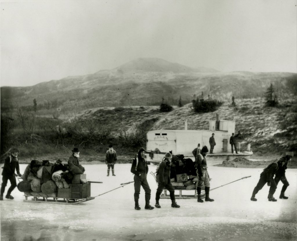 Black and white photo of people hauling sleds loaded with supplies down a frozen river.