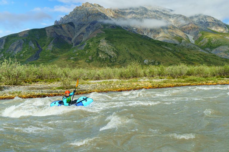 Packrafting and Aufeis in the Arctic National Wildlife Refuge Woman in blue packraft wearing orange helmet paddles on rough water on a river past a mountain