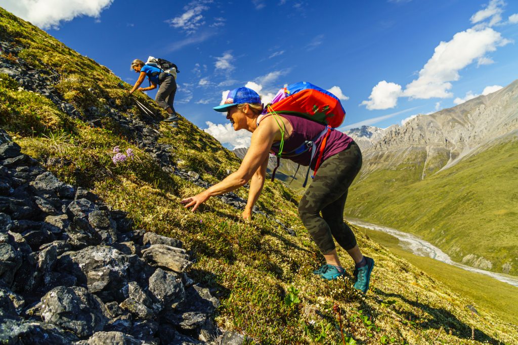 Two women scramble up a roughly 45-degree slope of rock and green tundra