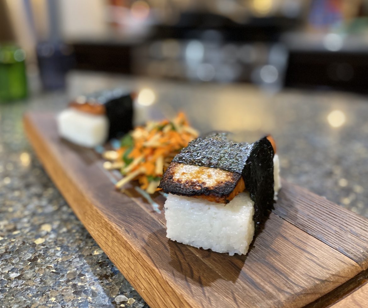 Two blocks of rice topped with fried salmon and wrapped in nori sit on either end of a wooden board with a small pile of slaw in the middle.