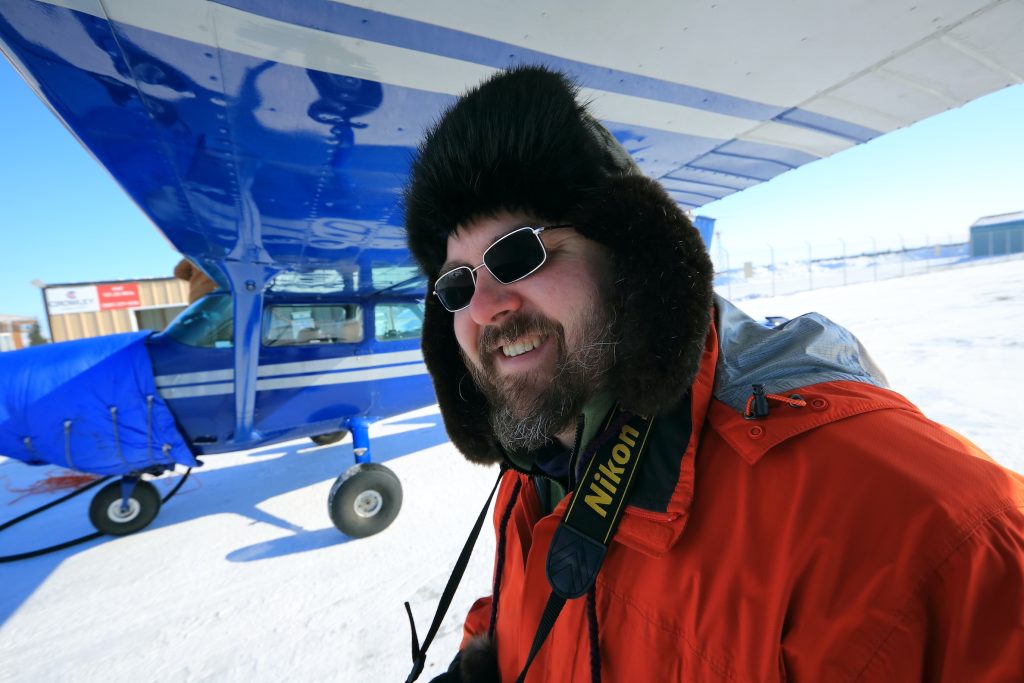 Bust shot of a man standing under plane wing in winter jacket and winter hat with a beard and camera around his neck