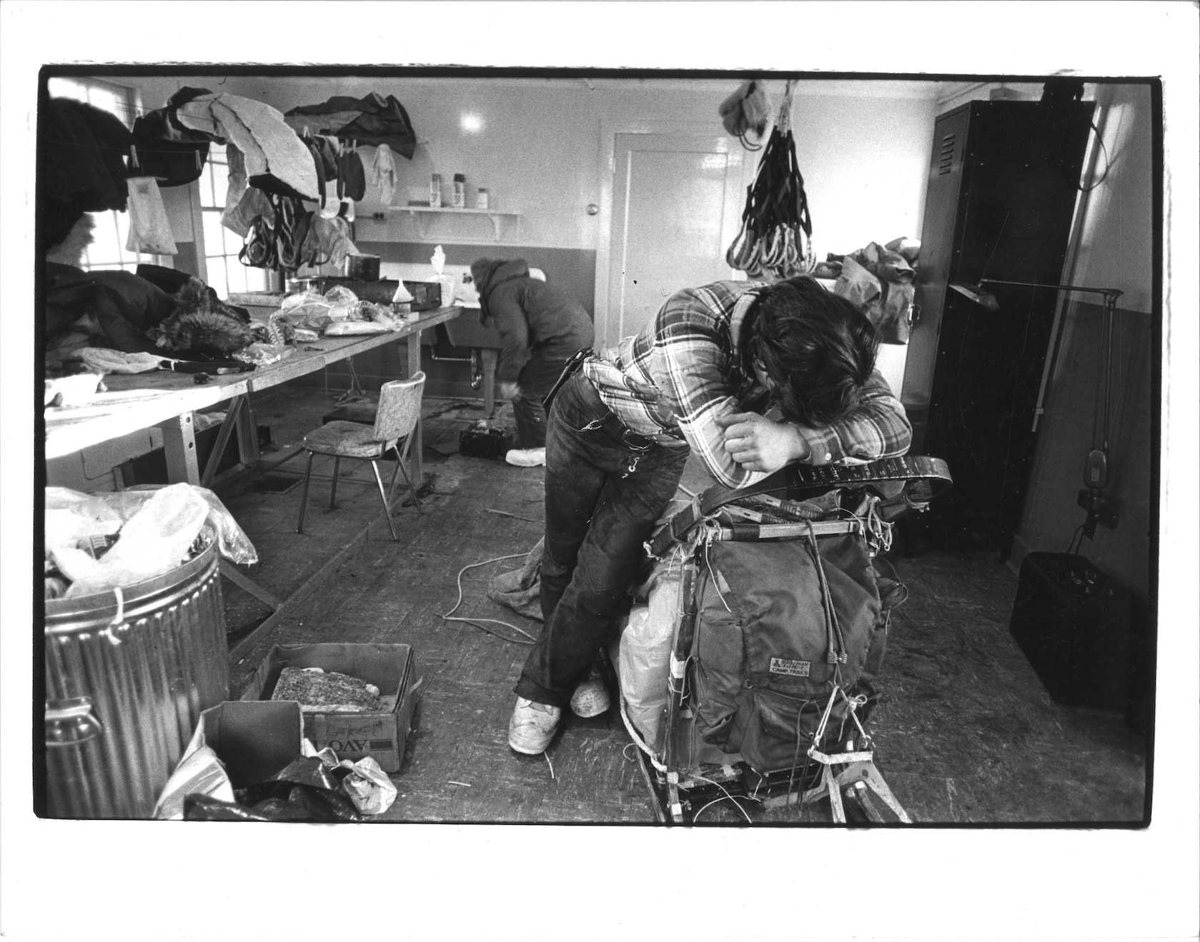 Musher leans on his sled and puts head in his folded arms. The room is strewn with gear. Black and white photo