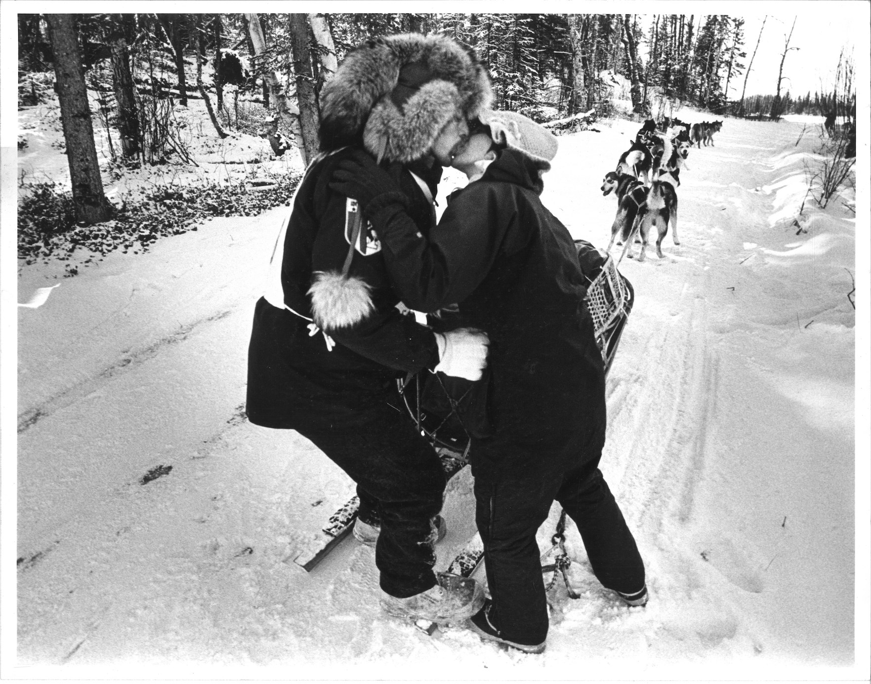 Musher stands on sled, dogs ready to run, and kisses his wife who is standing next to him on the snowy trail. Black and white photo.