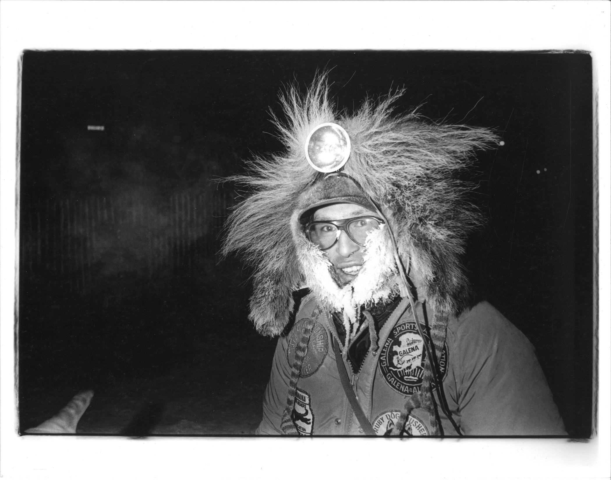 Man with big headlamp and hood covered in fur arrayed wildly, smiles at the camera in this black and white image.