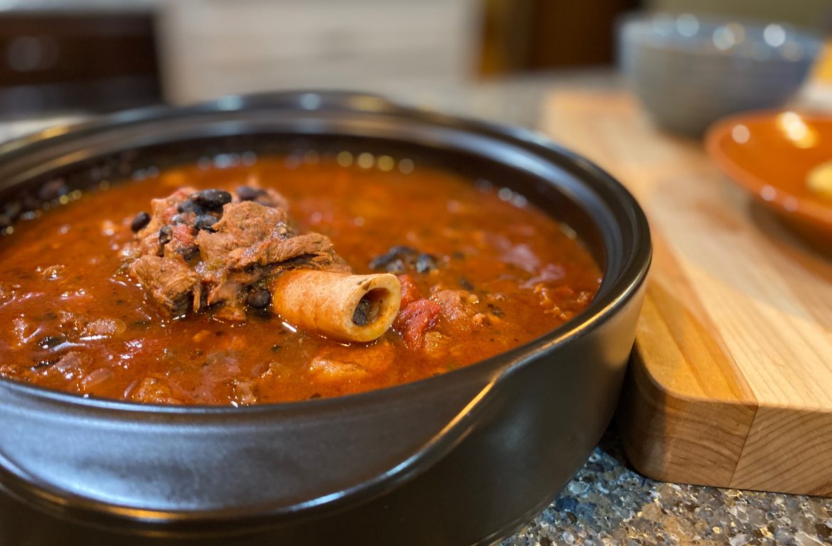 A bowl of red chili with a chunk of meat that still has the bone in.