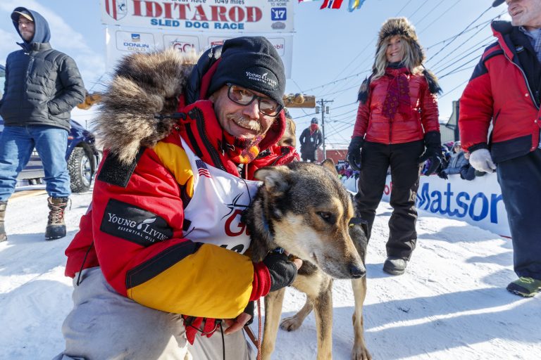Iditarod champion Mitch Seavey kneeling and hugging his dog under an Iditarod sign. He smiles for the camera.