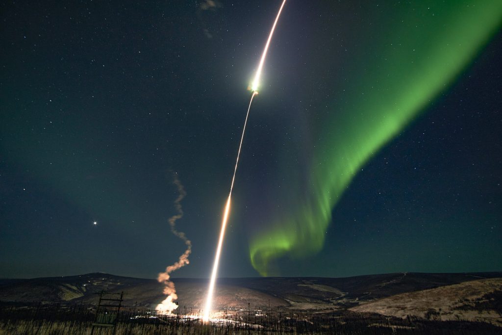 A rocket launches at night while a ribbon of aurora dances in the sky