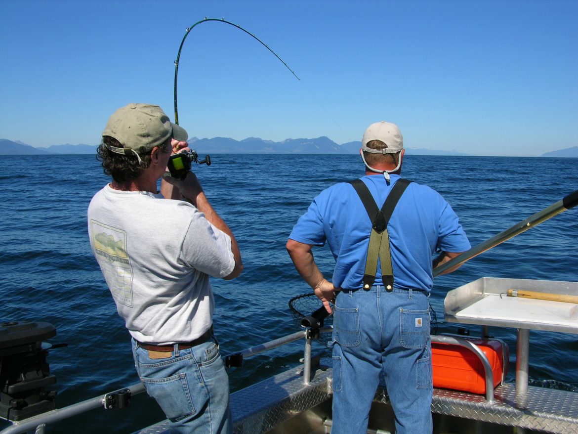 Nick Jans pulling back on a bent fishing rod, line in water, captain with his hands on hips watching for the fish to appear on the surface