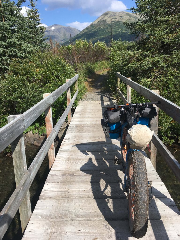 A bike loaded with bags sits on a narrow wooden bridge in Alaska's backcountry. Trees and mountains behind.