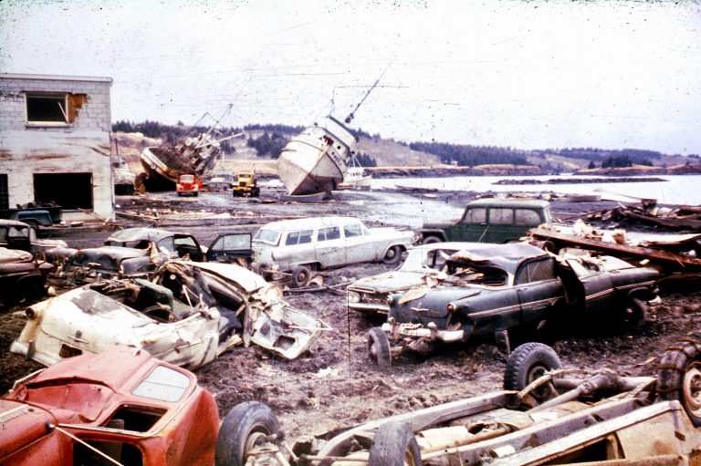 Cars, a fishing boat, building debris along a waterfront in old photograph