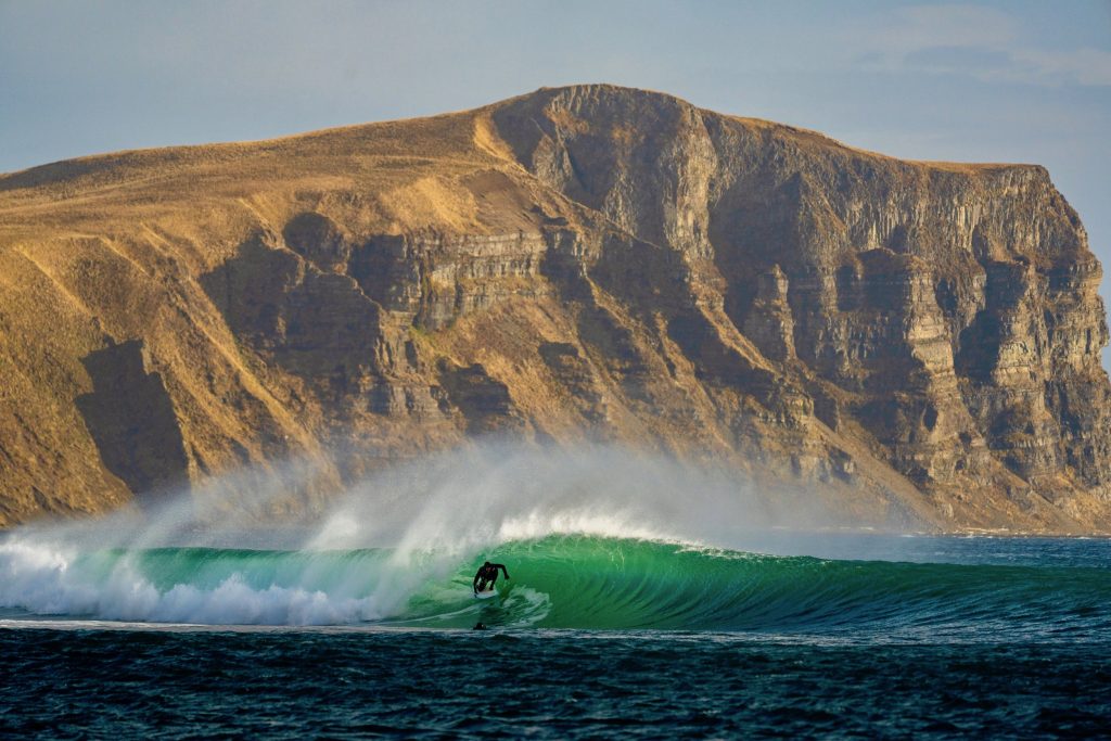 surfer in the barrel of a blue-green ocean wave. In the background is a tall mountain that slopes gradually on one side, and is cliffs on the other.