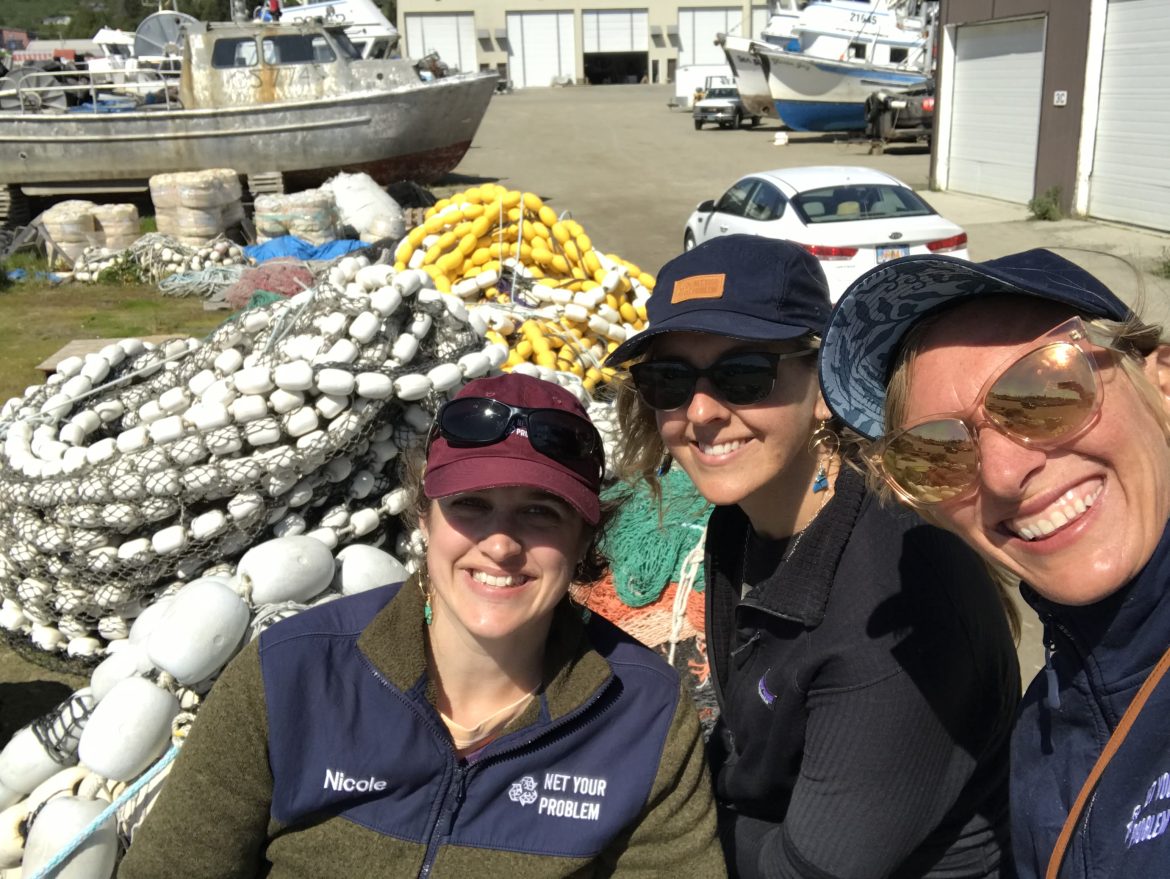 Recycling Alaska’s Plastic Fishing Nets Three women pose and smile in front of a pile of fishing nets