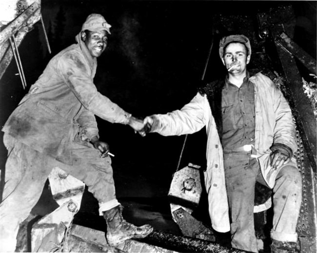 Black man and white man shake hands, both dressed warmly in this black and white photograph