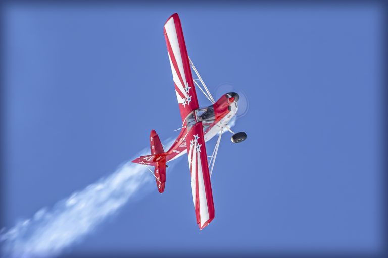 A red and white prop airplane flies on its side and white performance smoke streams out the back, highlighted against a blue sky