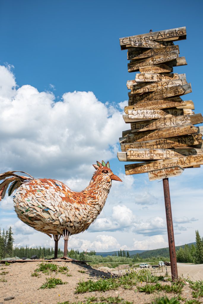 A giant chicken made of metal stands next to a cluttered sign pointing in many different directions