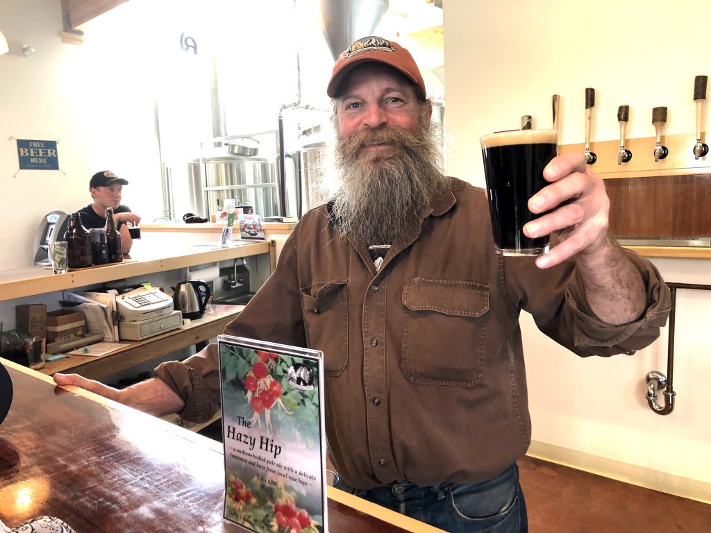 Bearded man standing behind wooden bar holds up a dark pint and smiles