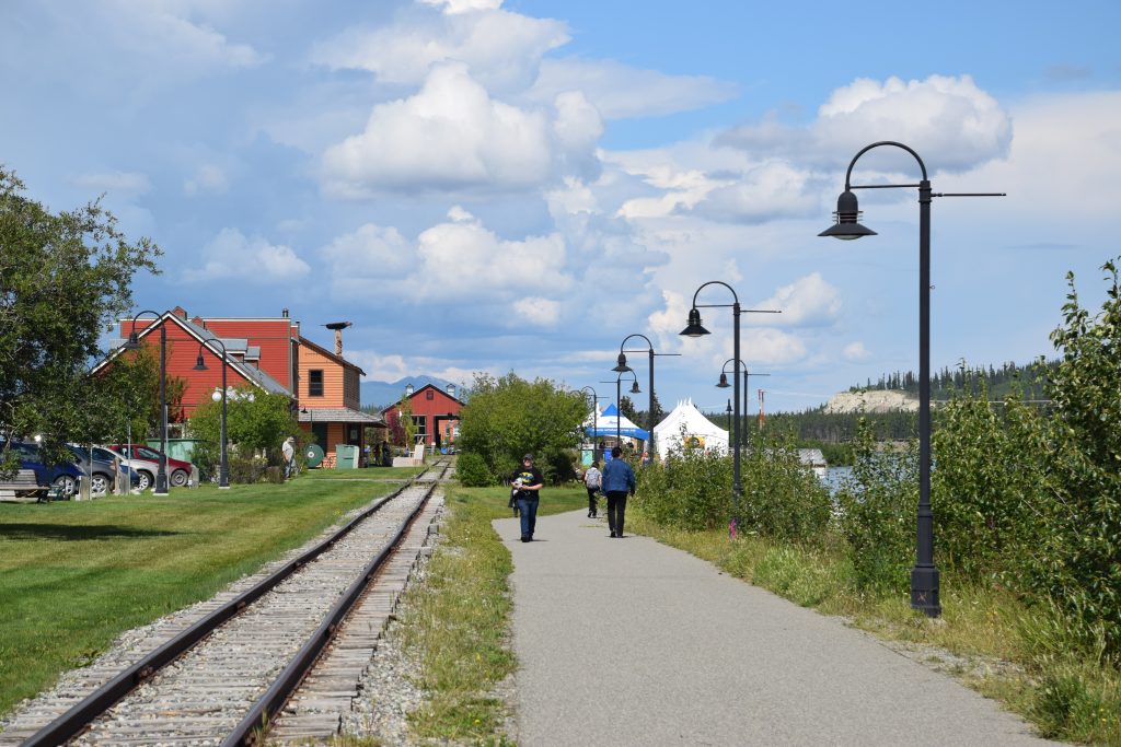 Train tracks next to a paved pathway that runs along a river. Buildings ahead and lampposts lining the walkway.