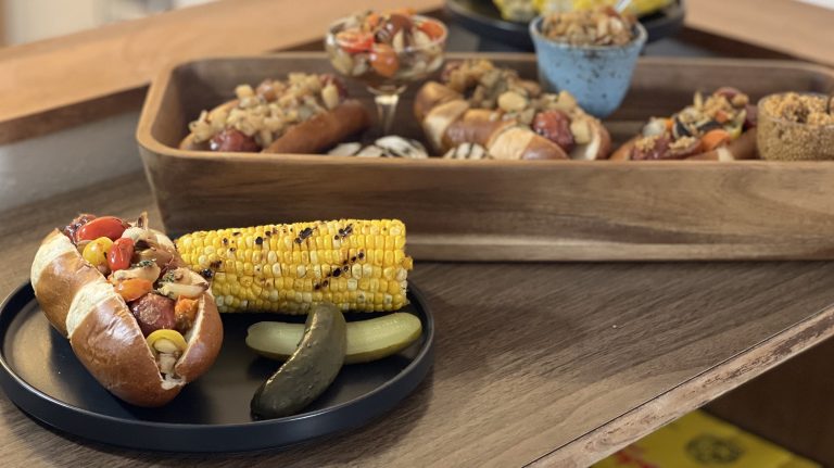 Sausage on a plate in pretzel bun, topped with colorful toppings, corn on the cob and pickles on the side.