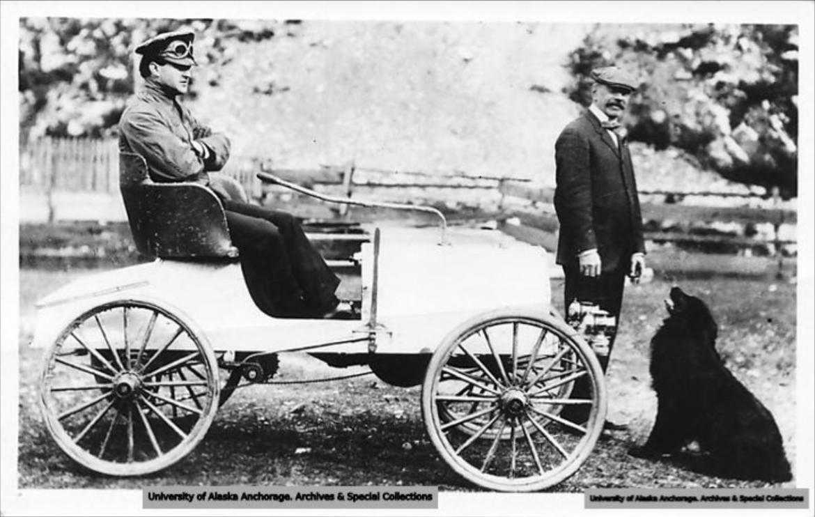 Black and white photo of a man sitting in an old car with round front and four wheels. Man stands in front with animal sitting at his legs.