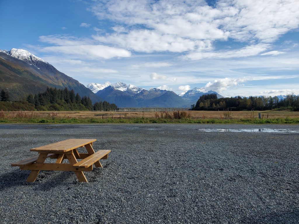 Wooden picnic table on gravel pad next to field with snow-capped mountains rising in distance