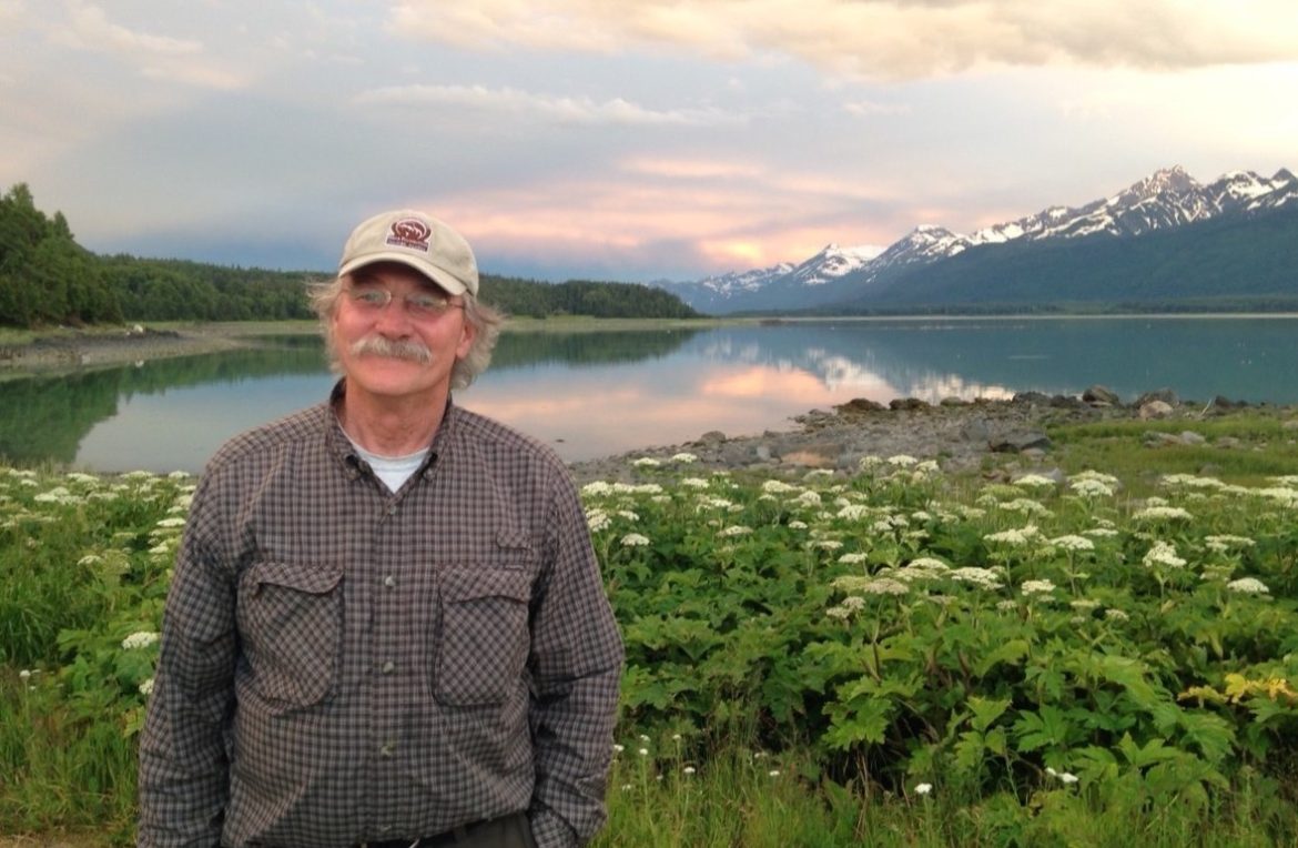 Dan Egolf, man with gray hair and ballcap, stands in front of foliage and a river