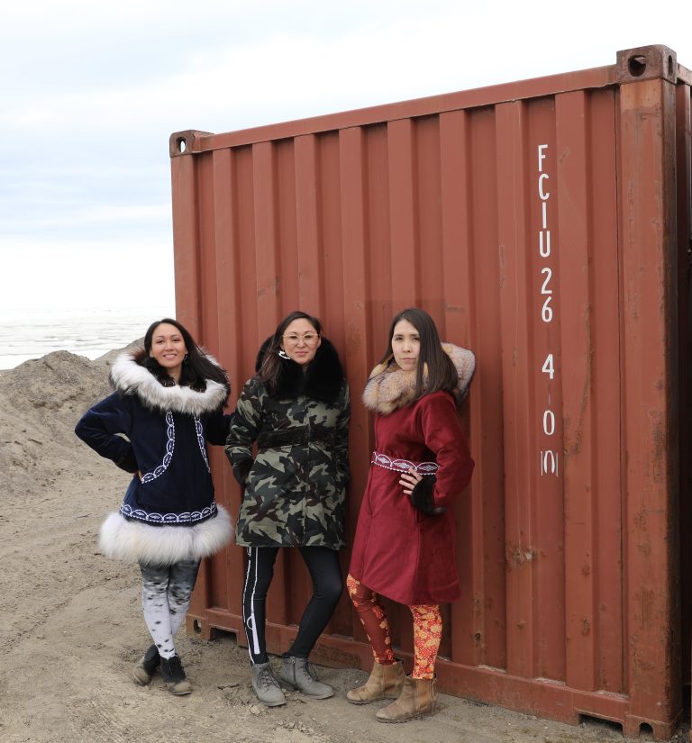 Three women pose in front of shipping container wearing stylized parkas and patterned leggings