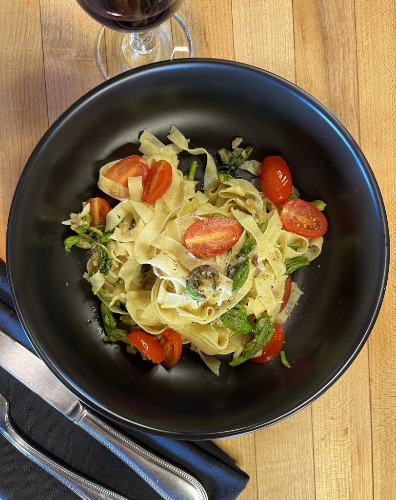 Image from above of black bowl with pasta, green shoots, and tomatoes inside